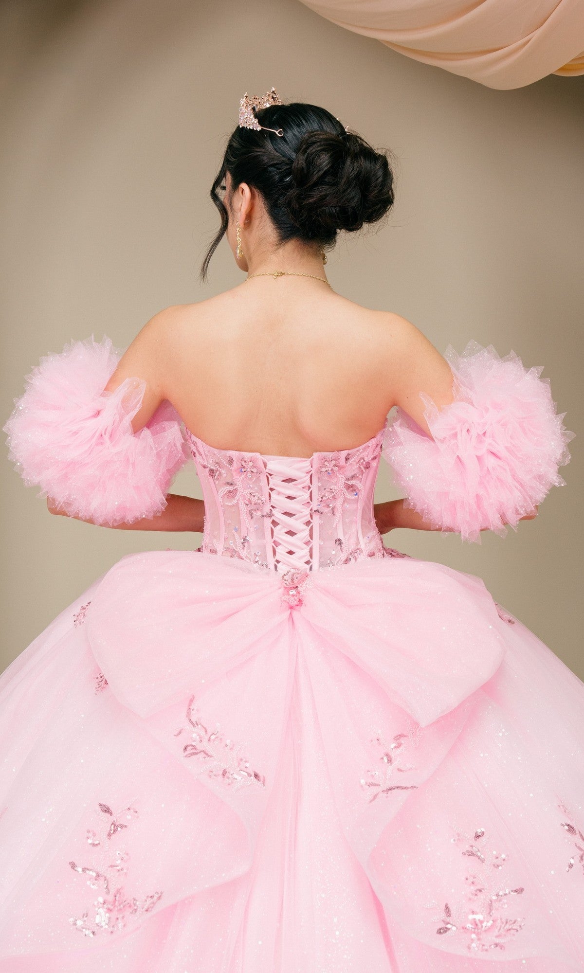 A person with dark hair in an updo is seen from the back wearing the Quinceanera Dress 8380J By Cinderella Couture, featuring a pink ball gown silhouette with a corset bodice, off-shoulder puff sleeves, and floral embellishments.